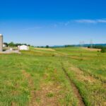 Wide rural farm scene with a silo, red-roofed barns, rolling green fields, and distant hills under a clear blue sky, with a dirt path leading toward the buildings.