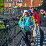 Group of hikers crosses fenced footbridge, historical brick townhomes in distance.