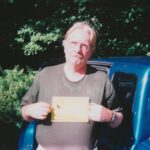 Man with glasses and a mustache standing outdoors beside a blue vehicle, holding a yellow plaque toward the camera.
