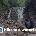 Two people sit on a rocky riverbank watching a multi‑tier waterfall in a forested setting.