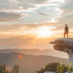 Person standing on a McAfee Knob cliff edge at sunrise, gazing over a valley and distant mountains below a colorful sky.