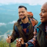 Two hikers with backpacks on a mountain trail, smiling as they overlook a valley below.