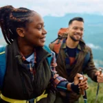 Two hikers with backpacks stand on a mountain trail, smiling as a scenic valley stretches behind them.