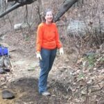 Woman in an orange sweatshirt and gloves standing on a dirt path in a wooded area with bare trees and fallen leaves.