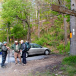 Four hikers with backpacks and trekking poles chat beside a parked silver car at an Appalachian Trail trailhead in a forest clearing.