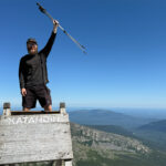 Hiker with a beard and cap raises trekking poles over a weathered Katahdin summit sign, blue sky and distant mountains behind him.