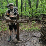 Hiker stands next to a wooden Mason-Dixon Line sign in a lush forest, Maryland/Pennsylvania border.