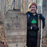 Smiling man in a brown knit cap and glasses stands in a forest beside a weathered wooden sign that reads 'Little Frog Wilderness' and 'Cherokee National Forest' with a large crosscut saw.