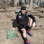 Hiker in black attire sits on a rocky trail with trekking poles and a backpack, near an Appalachian Trail plaque.