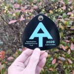Black Appalachian Trail marker tag with a large blue A/up-arrow and text 'Appalachian National Scenic Trail', '2026', and '2,197.9 miles' being held by a hand in front of colorful shrub leaves.