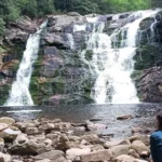 Two people sit on rocks by a waterfall in a rocky forested canyon.