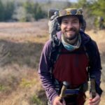 Smiling man with a beard hiking outdoors, wearing a large backpack, trekking poles, and goggles on his hat.