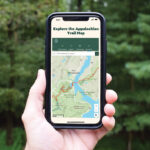 Hand holding a smartphone that shows an Appalachian Trail online map against a forest backdrop, with the header 'Explore the Appalachian Trail Map'.