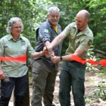 Three people in outdoor uniforms cut a red ribbon together at a woodland ceremony, celebrating a trail opening.