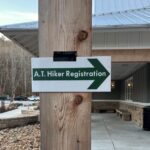 Green arrow sign reading 'A.T. Hiker Registration' mounted on a wooden post, directing hikers to the registration area near a building.