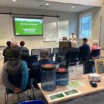 Indoor briefing session on the Appalachian Trail; presenter at a podium with a slide reading 'Start Smart at the Arch! Appalachian Trail Hiker Briefing'.