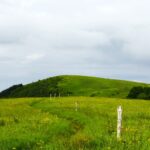 Pastoral meadow with a gentle green hill, a dirt path, and white fence posts under a cloudy sky.