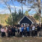 Group of diverse people posing outdoors in front of a small blue cabin, with trees and fallen leaves in autumn.
