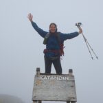 Hiker in a blue jacket stands atop a wooden summit sign with arms raised in celebration, trekking poles in hand (Katahdin).
