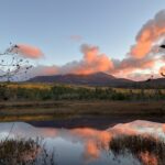 Sunset-lit mountain with pink clouds, autumn trees, and a calm reflective pond in the foreground.