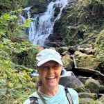 Smiling woman in a light blue shirt and white cap posing in front of a forest waterfall and rocks on a hike