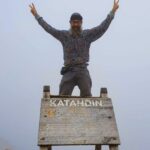 Man with a beard and glasses stands atop a wooden sign, arms raised in a peace sign, on Mt. Katahdin.