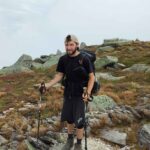 Hiker wearing black shirt and shorts with a backpack and trekking poles on a rocky alpine trail.105?