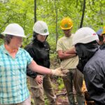 Group of people in hard hats and outdoor gear stand in a forest, examining a muddy rock or sample together during a field workshop.
