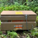 Brown KNAAK (Jobmaster) metal storage box with a chain lock, set among ferns and trees in a forest foreground.