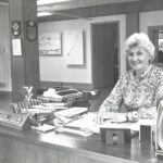 Woman stands at office desk, smiling at the camera; nameplate reads 'Jean Cashin' (Recreation Specialist).