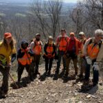 Group of hikers in orange safety vests on a rocky trail, with leafless trees and a valley view in the distance.