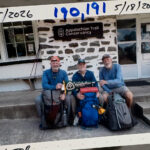 Three hikers with large backpacks sit on steps outside a stone building, holding an Appalachian Trail sign.