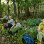Five people rest along a forest trail, with backpacks; a person in a bright yellow shirt sits on a rock to the right while others sit or stand on the left. An orange measuring wheel lies on the ground near a tree.