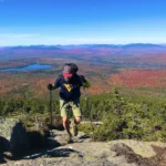 Hiker with pack and trekking poles climbs a rocky summit along the Appalachian Trail, vast autumn landscape in the distance.