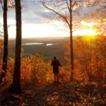 Hiker surrounded by fall foliage overlooks a mountain vista, sun rising over the horizon.