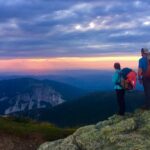 Two hikers with packs stand on a rocky outcrop, overlooking a distant mountain range at sunset.