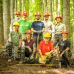 Nine members of the Konnarock Crew pose in hardhats along the Appalachian Trail in a forested setting, with some holding hand tools.