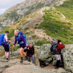 A group of hikers climbs a rocky section of the Appalachian Trail, with more hikers visible along the distant ridgeline.