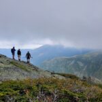 Three hikers stand on a rocky slope of Mt Adams, looking out at large, distant mountains obscured by clouds.