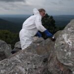 Volunteer wearing coveralls and gloves scrubs graffiti off a rocky mountain surface with a brush.